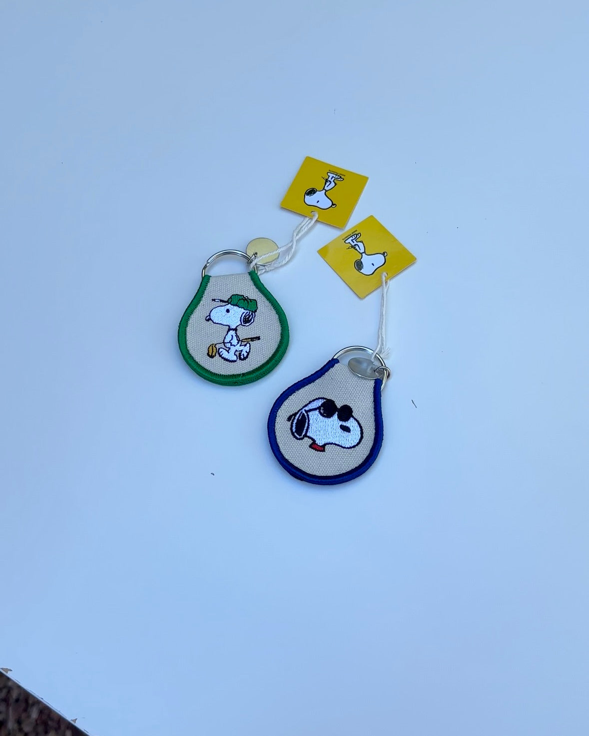 Two Snoopy Peanuts embroidered keychains, one in green with tennis Snoopy and one in blue with sunglasses Snoopy, displayed on a white background.