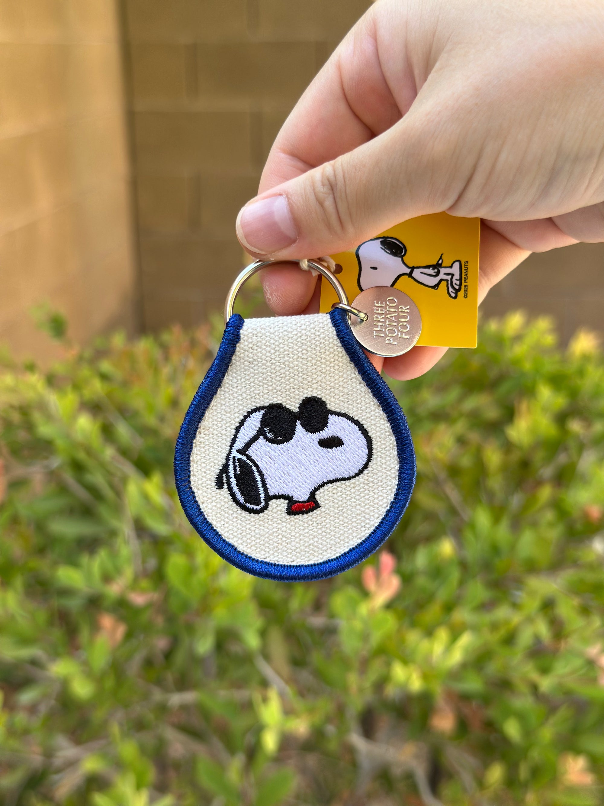 Close-up of Snoopy Peanuts embroidered keychain with blue trim featuring sunglasses Snoopy, held outdoors with greenery in background.