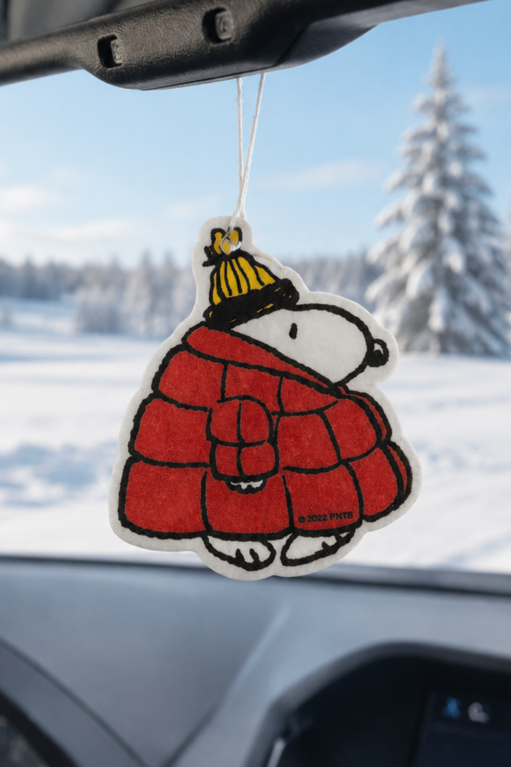 Snoopy car air freshener wearing a red puffer jacket and yellow beanie with Woodstock on top, hanging in a car with a snowy pine forest outside.