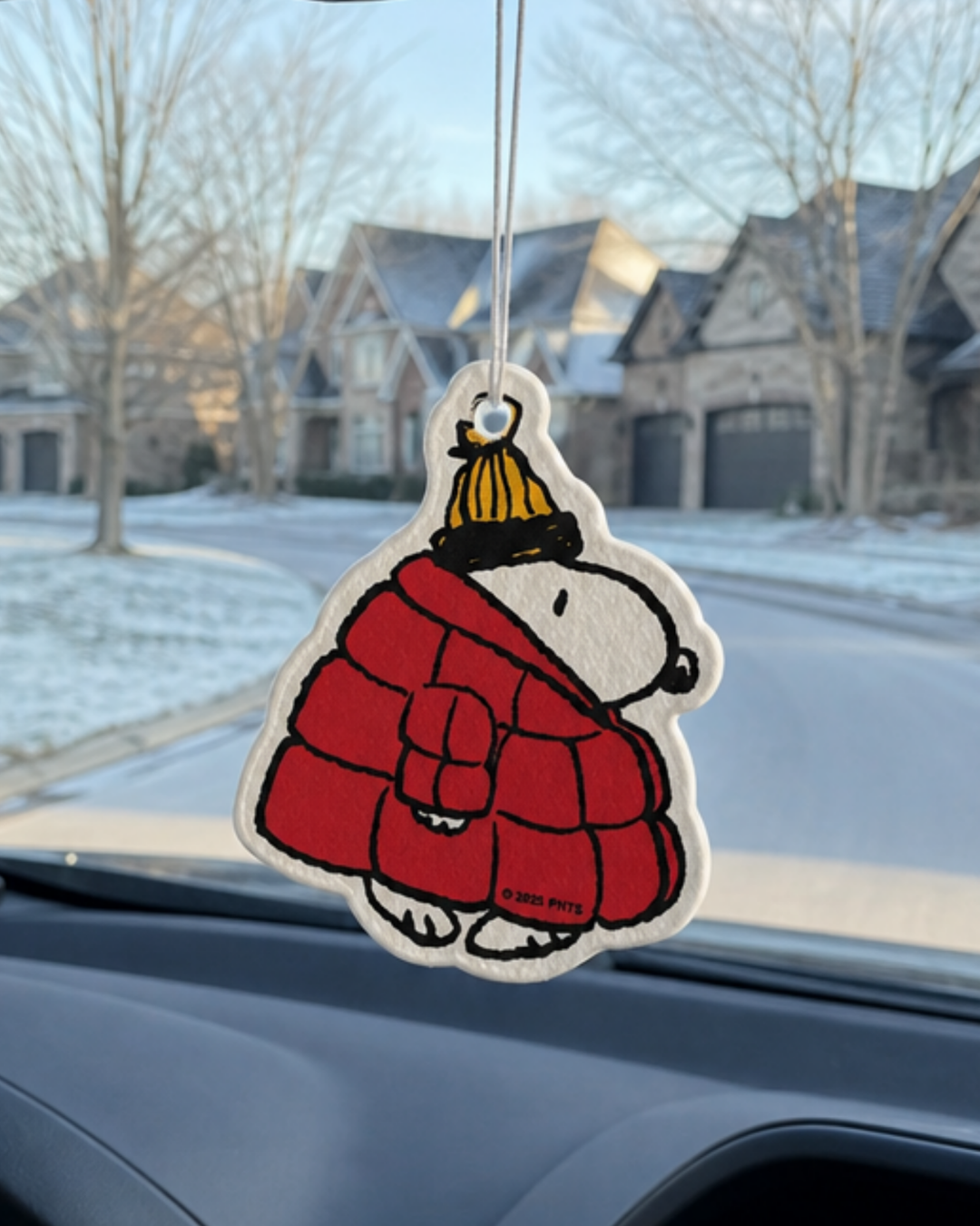 Snoopy air freshener in a red puffer jacket and yellow beanie with Woodstock perched on top, hanging from a car mirror on a snowy suburban street.
