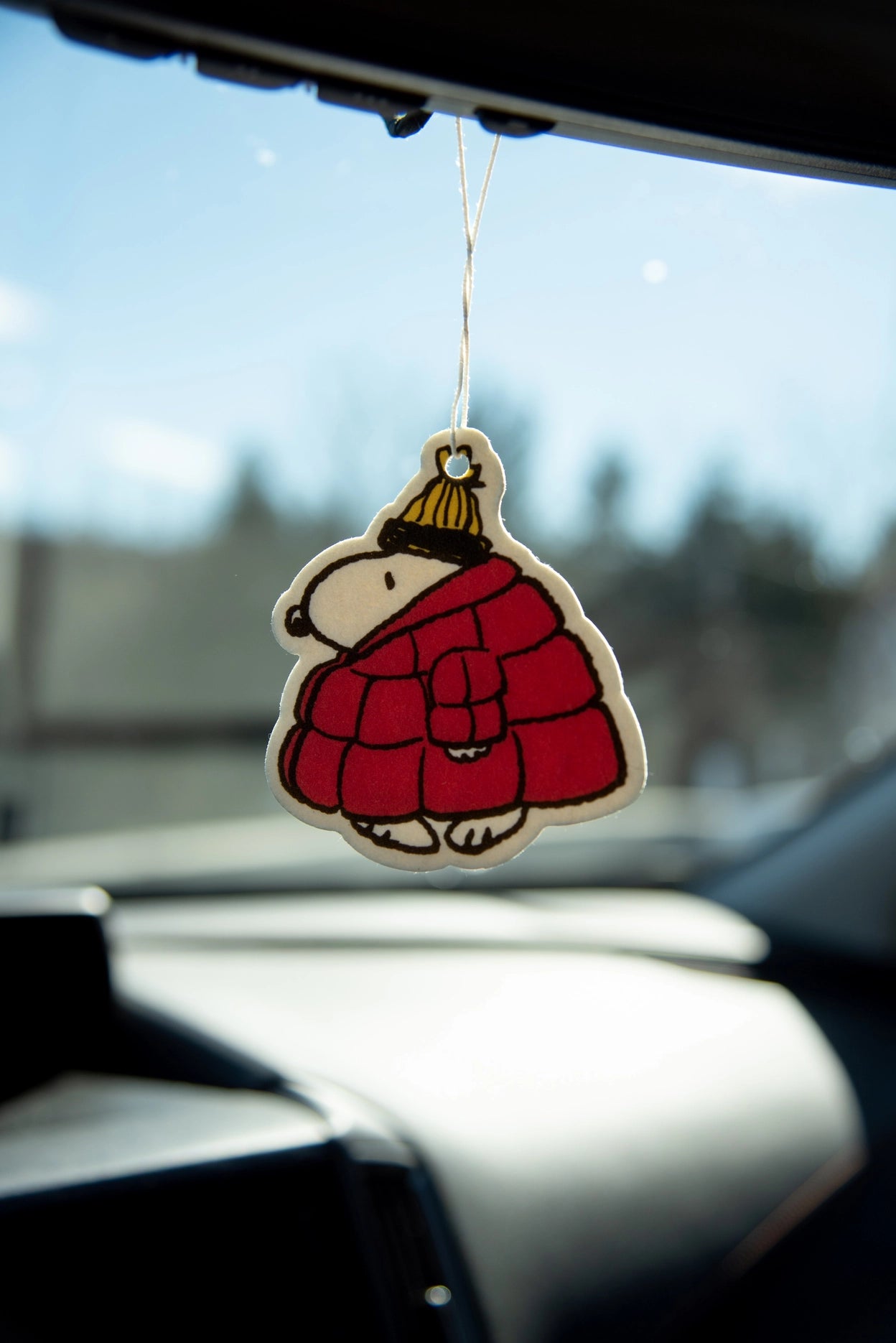 Snoopy puffer jacket air freshener hanging from a car mirror with blurred outdoor background.