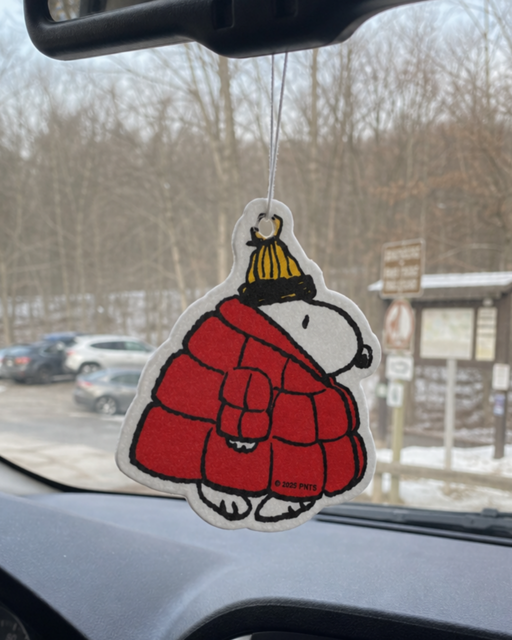Snoopy air freshener in a red puffer jacket and yellow beanie, hanging from a car mirror near a snowy trailhead parking area.