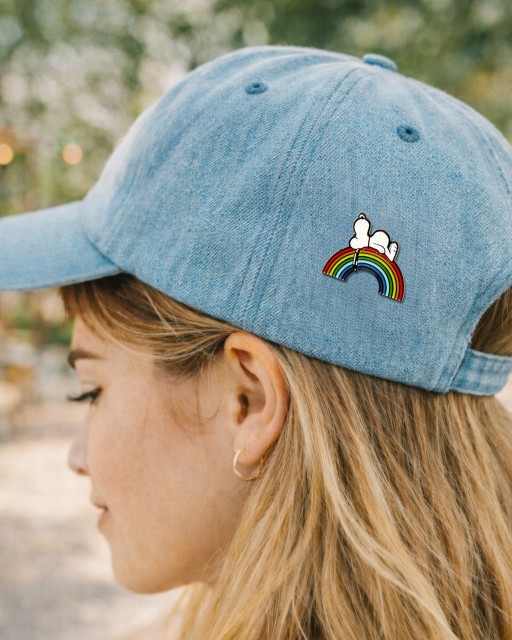 Snoopy rainbow enamel pin on a blue baseball cap, cute Peanuts accessory for hats, jackets, and enamel pin collectors.