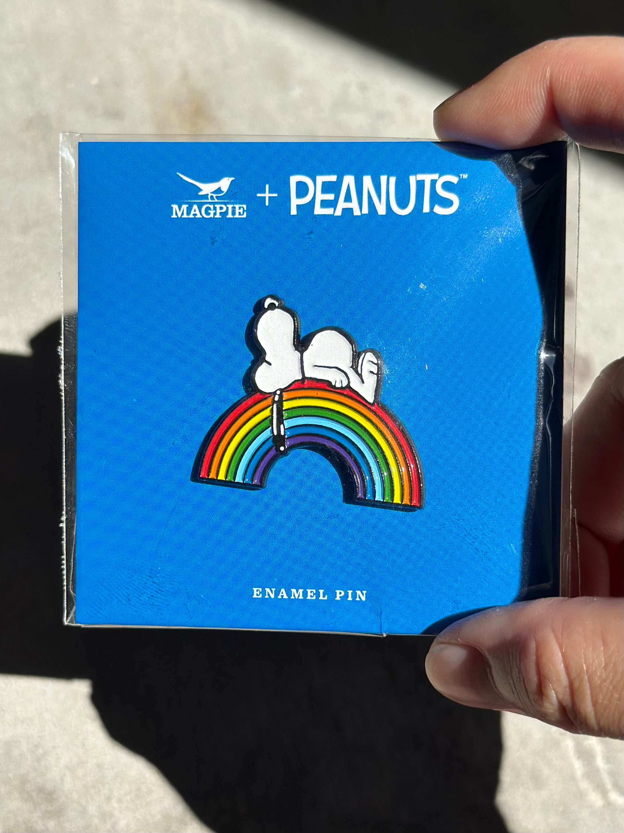 Snoopy enamel pin lounging on rainbow, displayed on Magpie + Peanuts backing card.