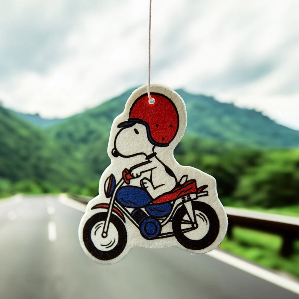 Snoopy motorcycle air freshener hanging in front of a scenic road view. Sandalwood-scented car decor for Peanuts lovers and road trip vibes.