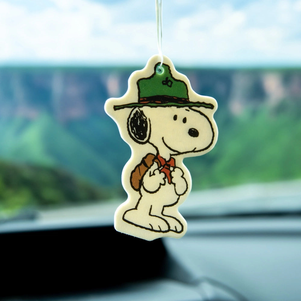 Scout Snoopy air freshener hanging from a car mirror with green cliffs in the background. Woodsy Peanuts-themed accessory for road trip vibes.