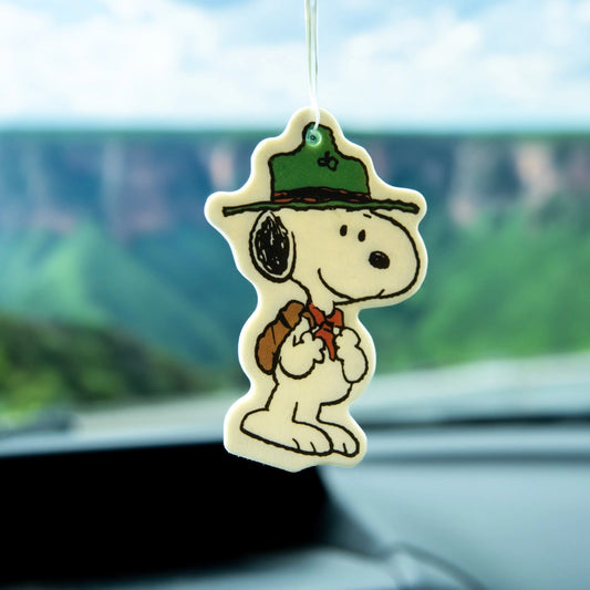 Scout Snoopy air freshener hanging from a car mirror with green cliffs in the background. Woodsy Peanuts-themed accessory for road trip vibes.