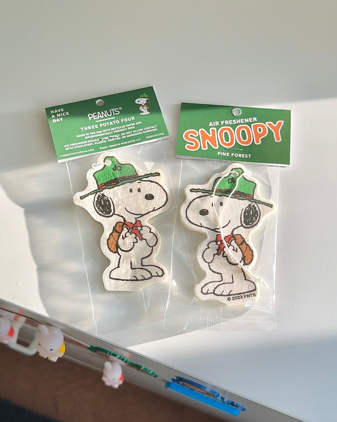 Snoopy Scout air fresheners with vintage-style packaging, featuring a green hat and red neckerchief, shown side by side on a white surface.