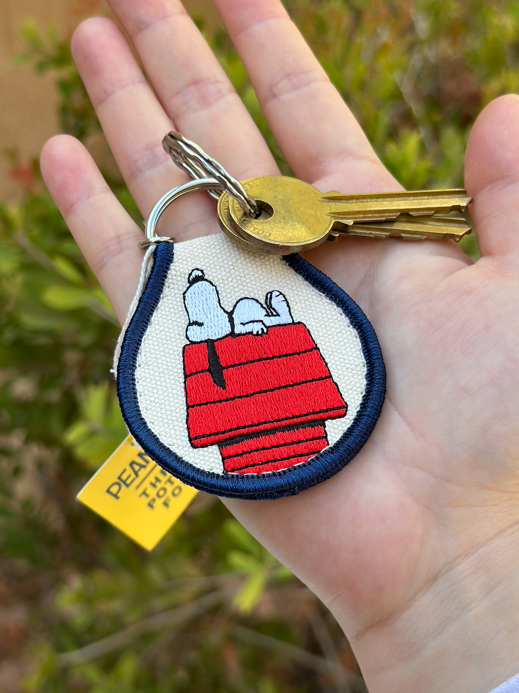 Hand holding Snoopy Peanuts embroidered keychain with Snoopy sleeping on his red doghouse, navy trim, and silver split ring.