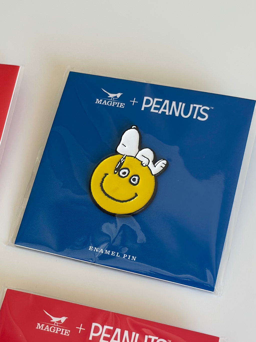 Snoopy enamel pin on yellow smiley face, displayed flat on Magpie + Peanuts collector card.