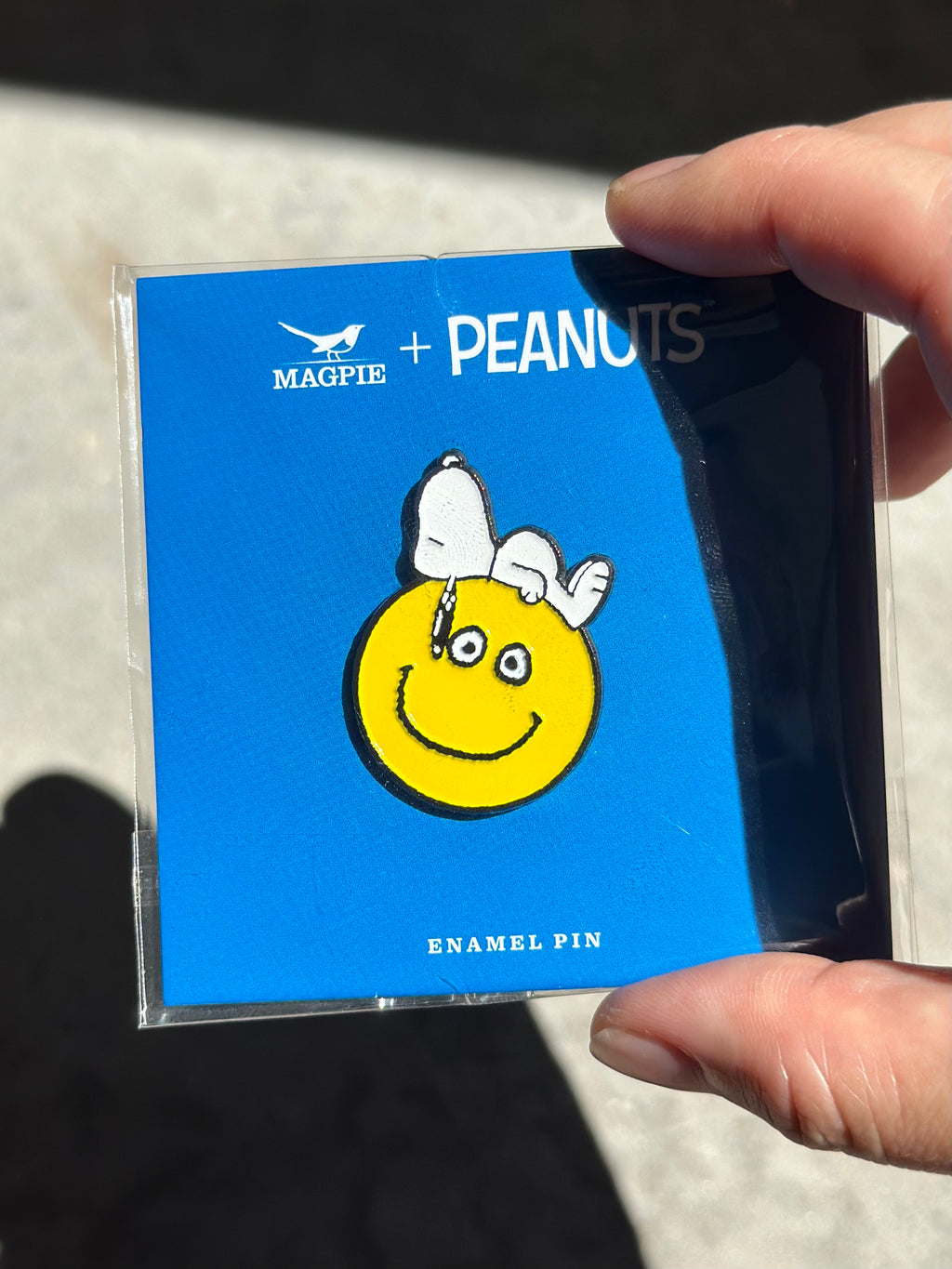 Snoopy enamel pin on yellow smiley face, held in sunlight with Magpie + Peanuts backing card.