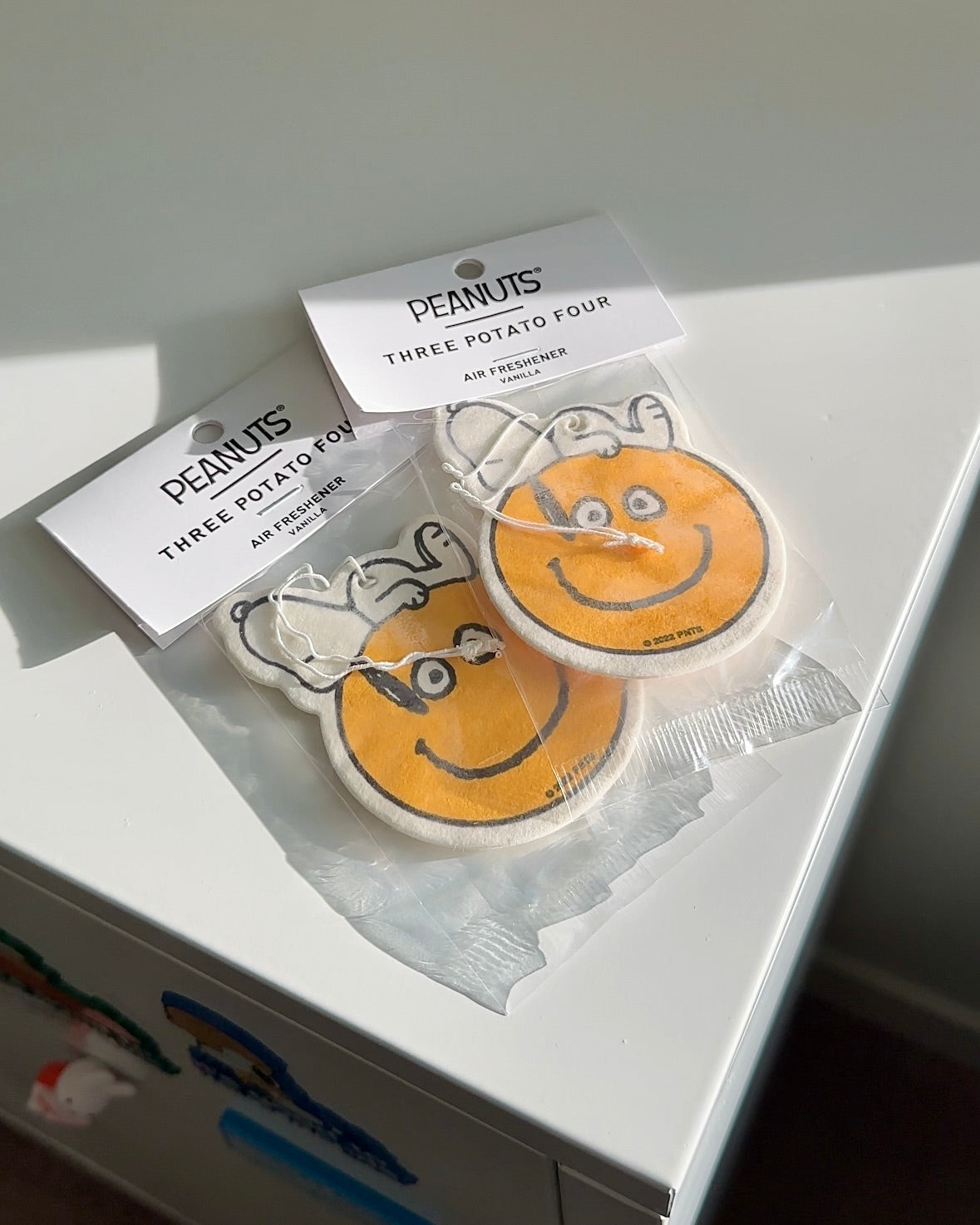 Two yellow smiley face air fresheners featuring a relaxed Snoopy lying on top, displayed in natural sunlight on a white surface.