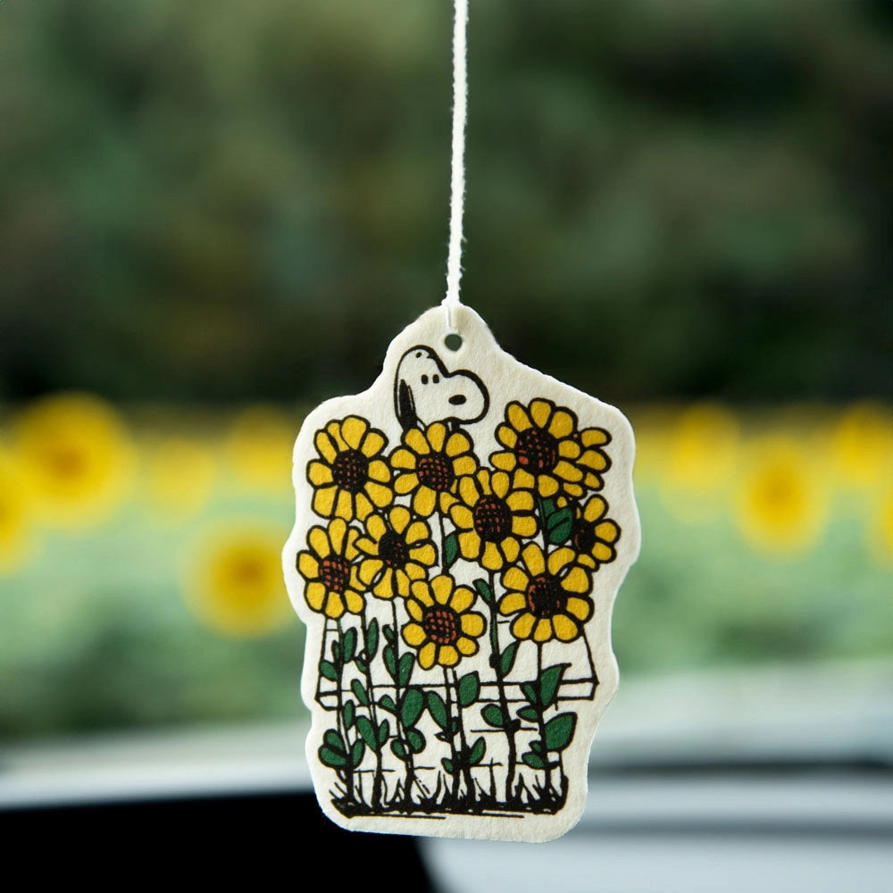 Snoopy air freshener with sunflower design hanging in front of a car window. Yellow flowers and lavender scent give cozy, summer-ready energy.