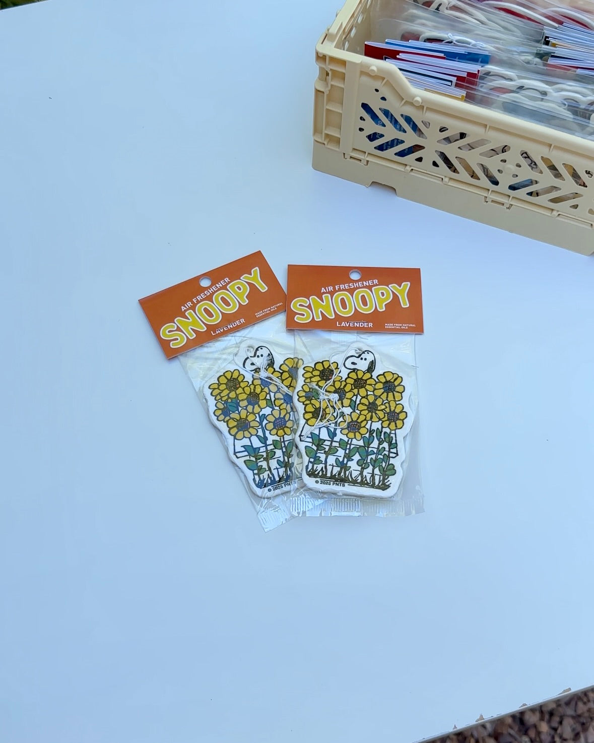 Two packaged Snoopy Sunflower lavender-scented air fresheners on a white table with a beige crate in the background.