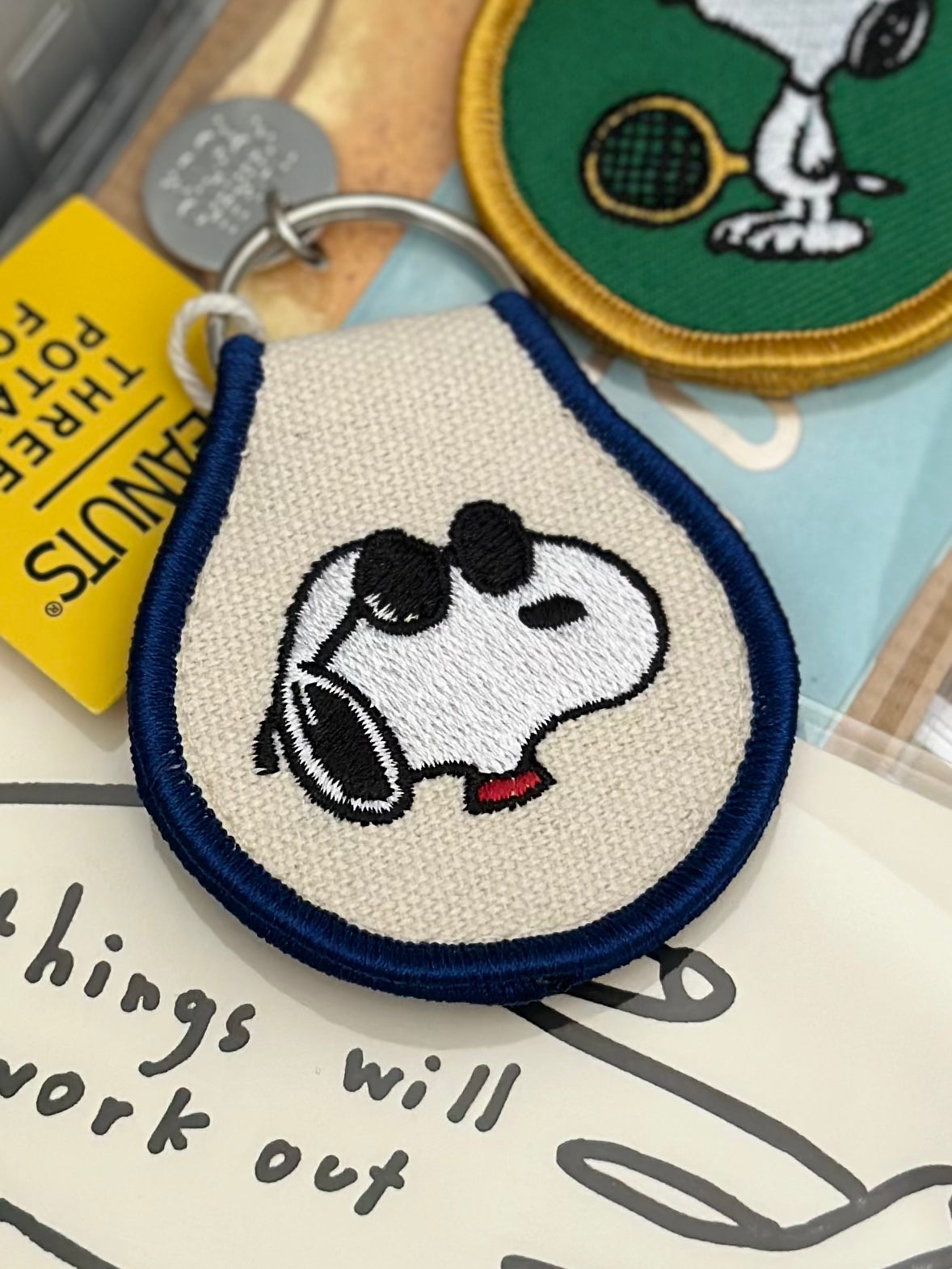 Embroidered Snoopy face keychain with sunglasses on cream fabric and blue trim.