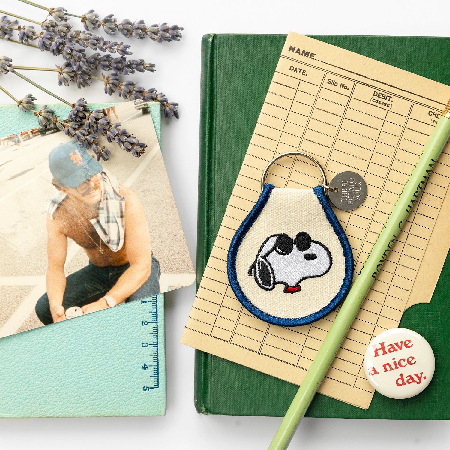 Flat lay featuring an embroidered Snoopy keychain with blue trim, styled with vintage photo, dried lavender, and a “Have a nice day” button. A retro-inspired Peanuts keychain that brings personality to your bag or keys.
