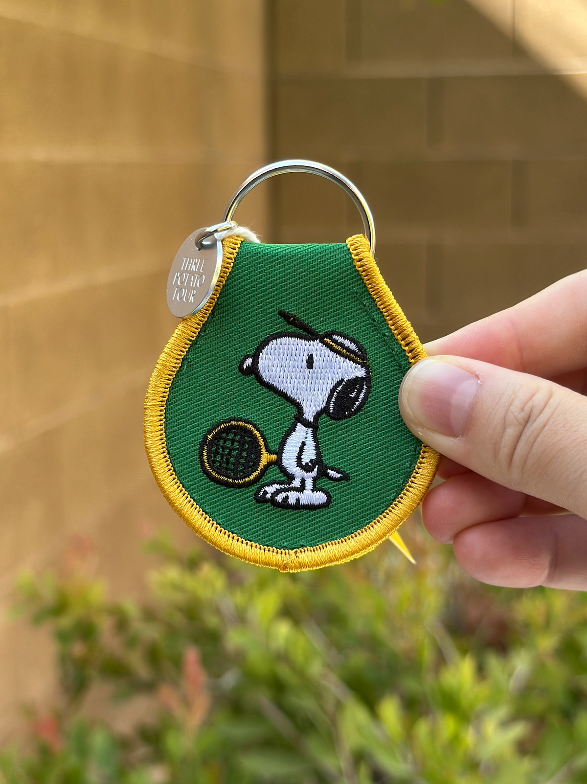 Snoopy Peanuts embroidered tennis keychain with yellow trim, held outdoors with greenery background.