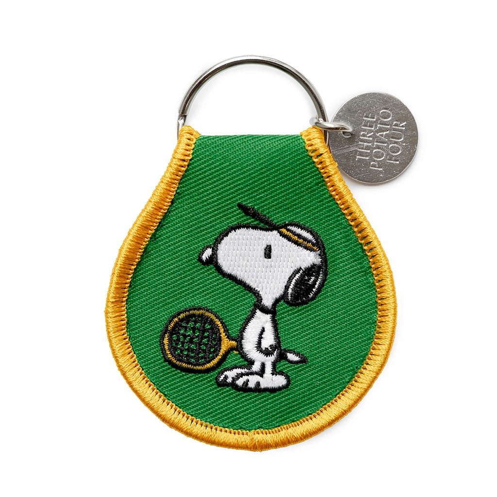 Close-up of embroidered Snoopy keychain with green background and yellow trim. Features Snoopy in a tennis visor holding a racket, attached to a silver key ring with Three Potato Four branding tag.
