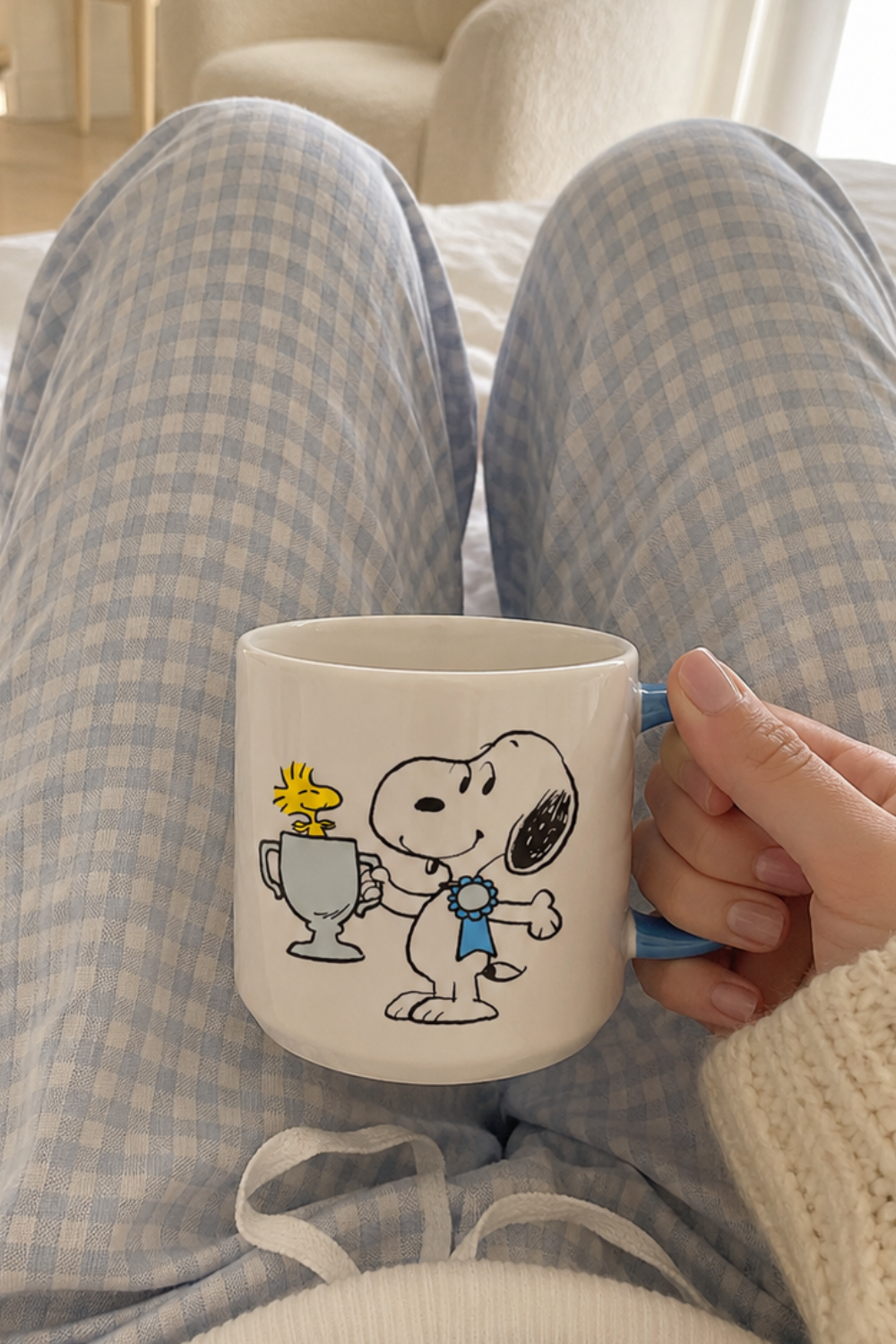 Snoopy Top Dog ceramic mug held above blue gingham pajamas in a bright and cozy bedroom setting.
