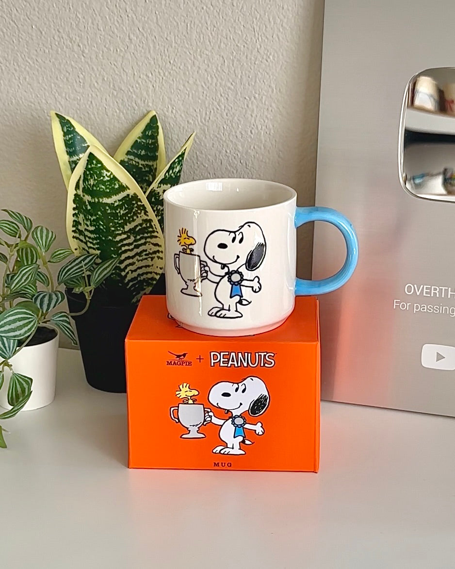 Snoopy Top Dog mug placed on its orange box, surrounded by indoor plants and a YouTube plaque in the background