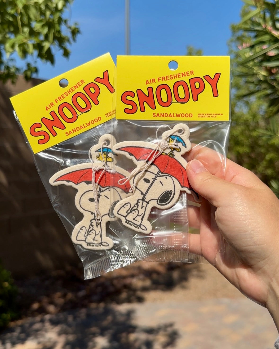 Hand holding two packaged Snoopy Umbrella sandalwood-scented air fresheners against a sunny blue sky with greenery.
