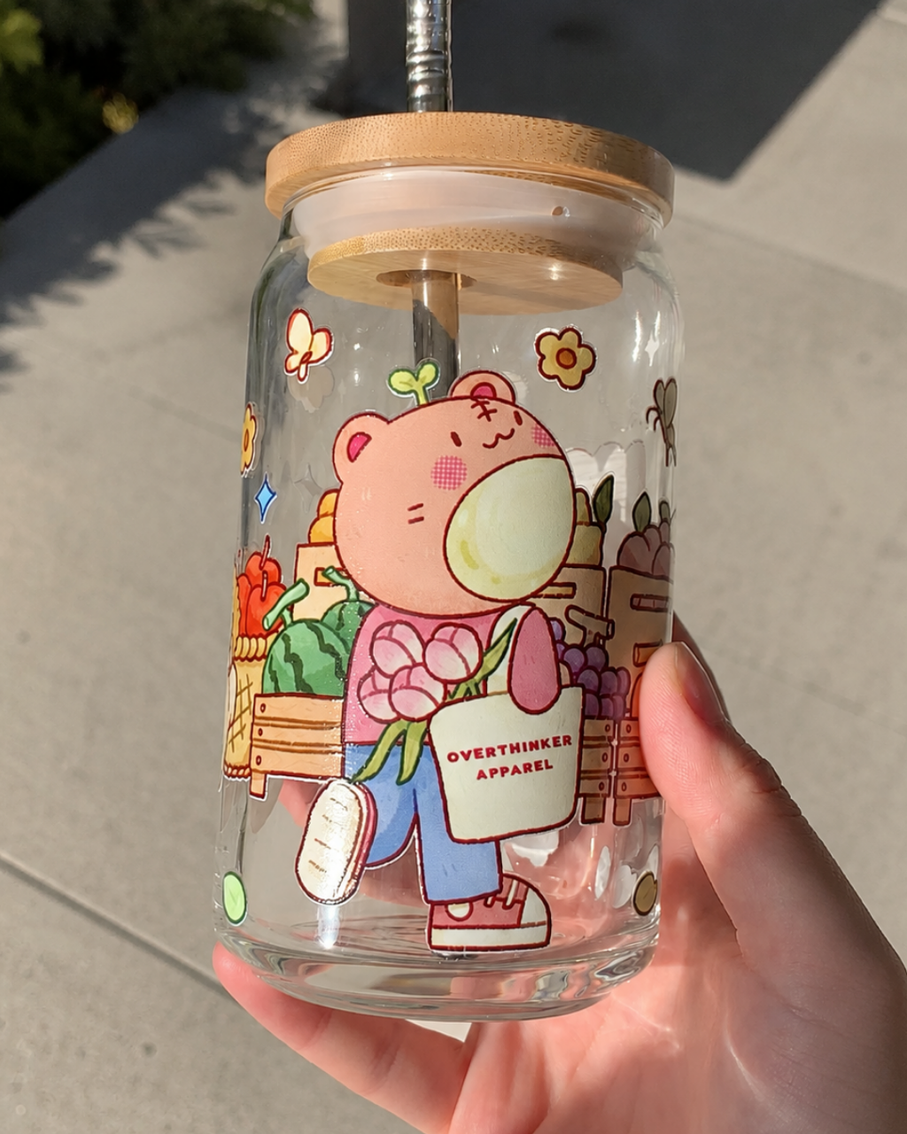 A clear glass straw tumbler with a bamboo lid is held in a hand outdoors. The tumbler features a colorful cartoon illustration of a pink bear holding a bag that reads 'OVERTHINKER APPAREL'.