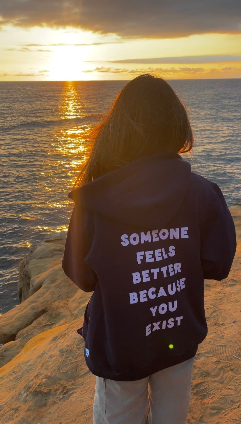 Navy zip-up hoodie with the affirmation “someone feels better because you exist” printed on the back, worn outdoors at sunset by the ocean, calm and emotionally supportive clothing.