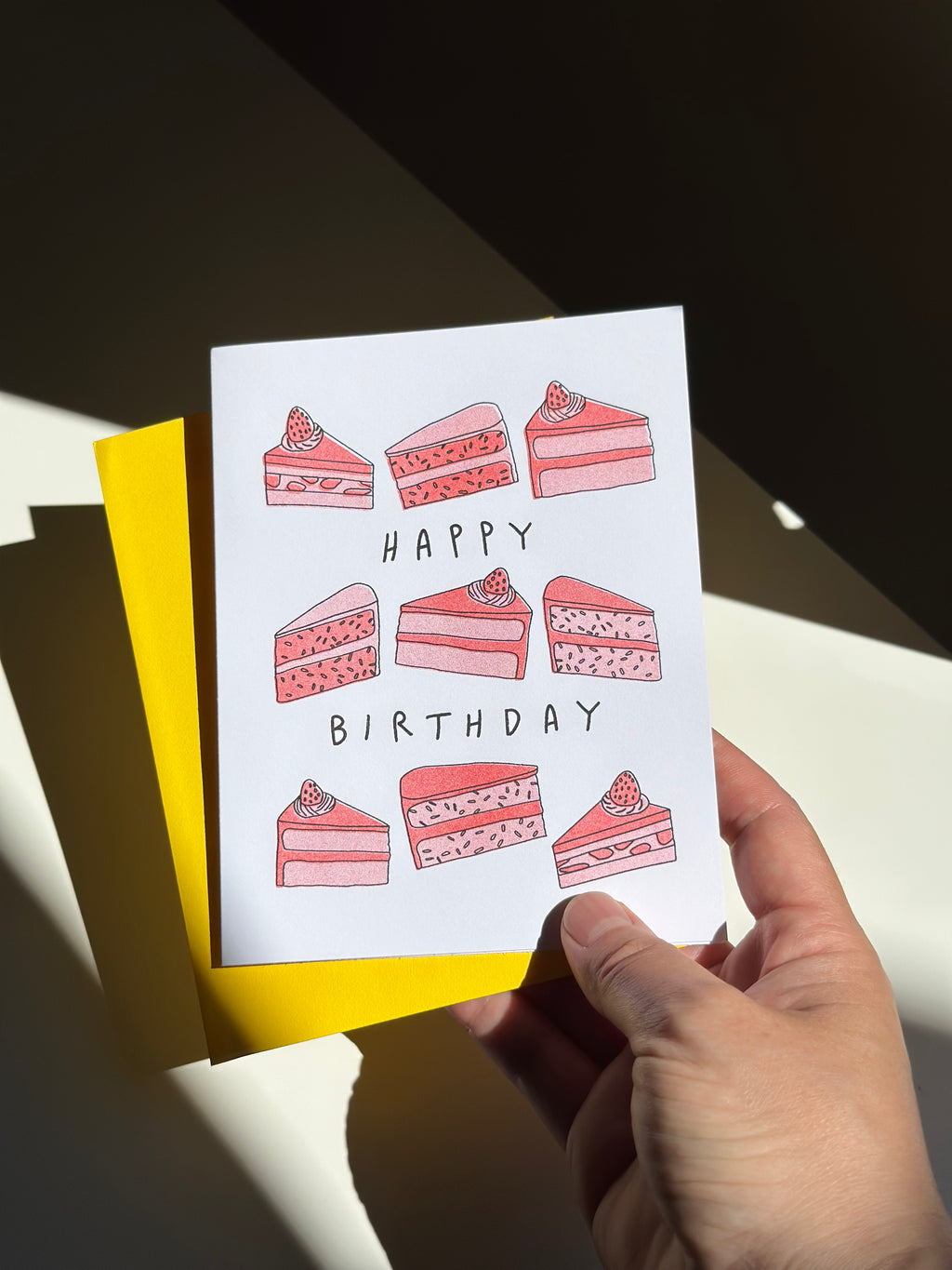 Happy birthday card with hand-drawn slices of pink strawberry cake with strawberries on top, arranged around the words ‘Happy Birthday,’ paired with bright yellow envelope.