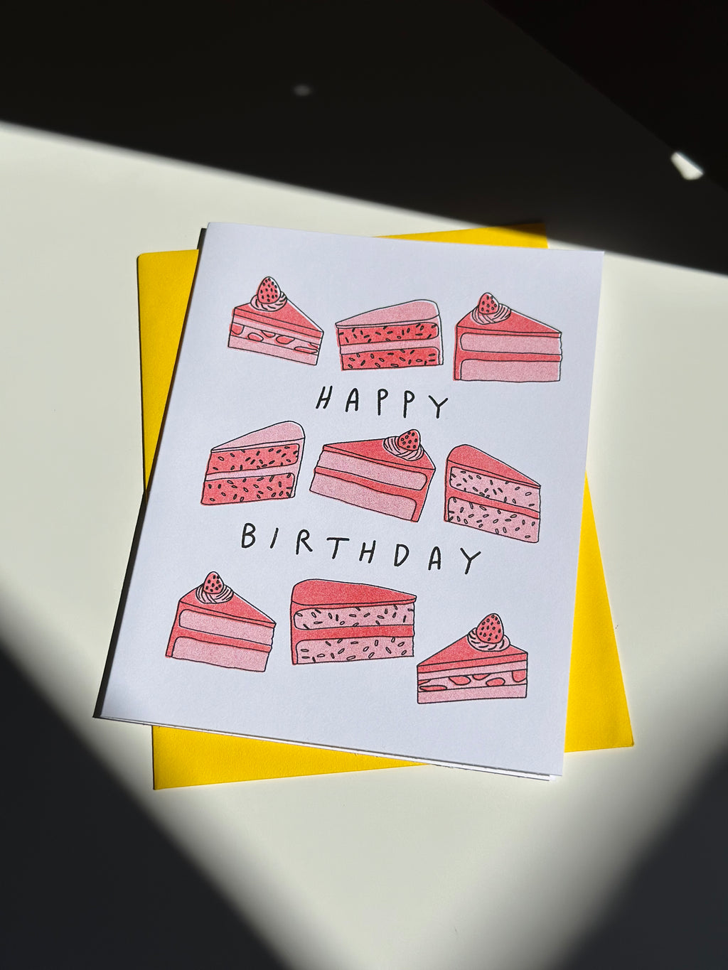 Happy birthday card with hand-drawn slices of pink strawberry cake with strawberries on top, arranged around the words ‘Happy Birthday,’ paired with bright yellow envelope.