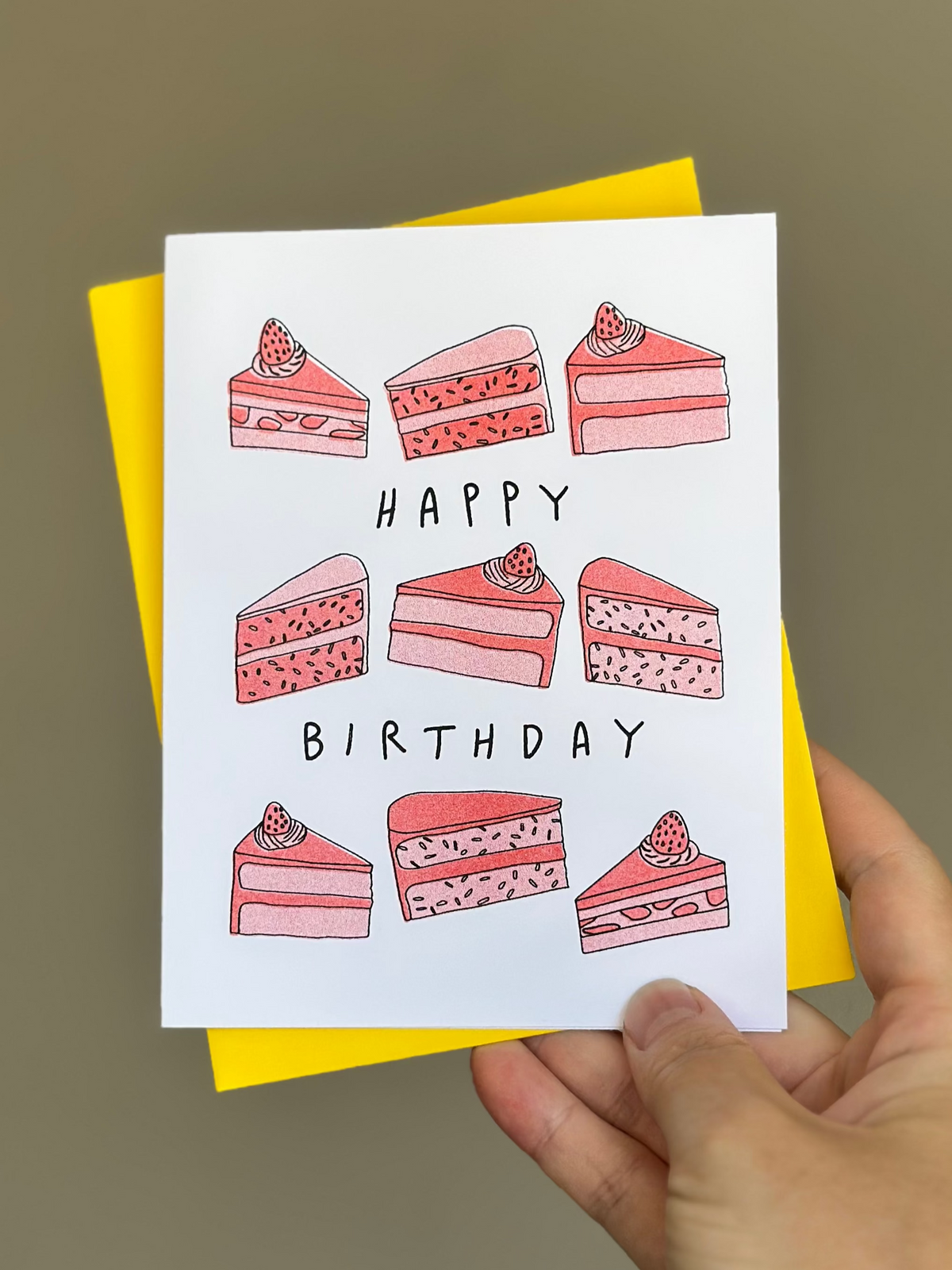 Happy birthday card with hand-drawn slices of pink strawberry cake with strawberries on top, arranged around the words ‘Happy Birthday,’ paired with bright yellow envelope.