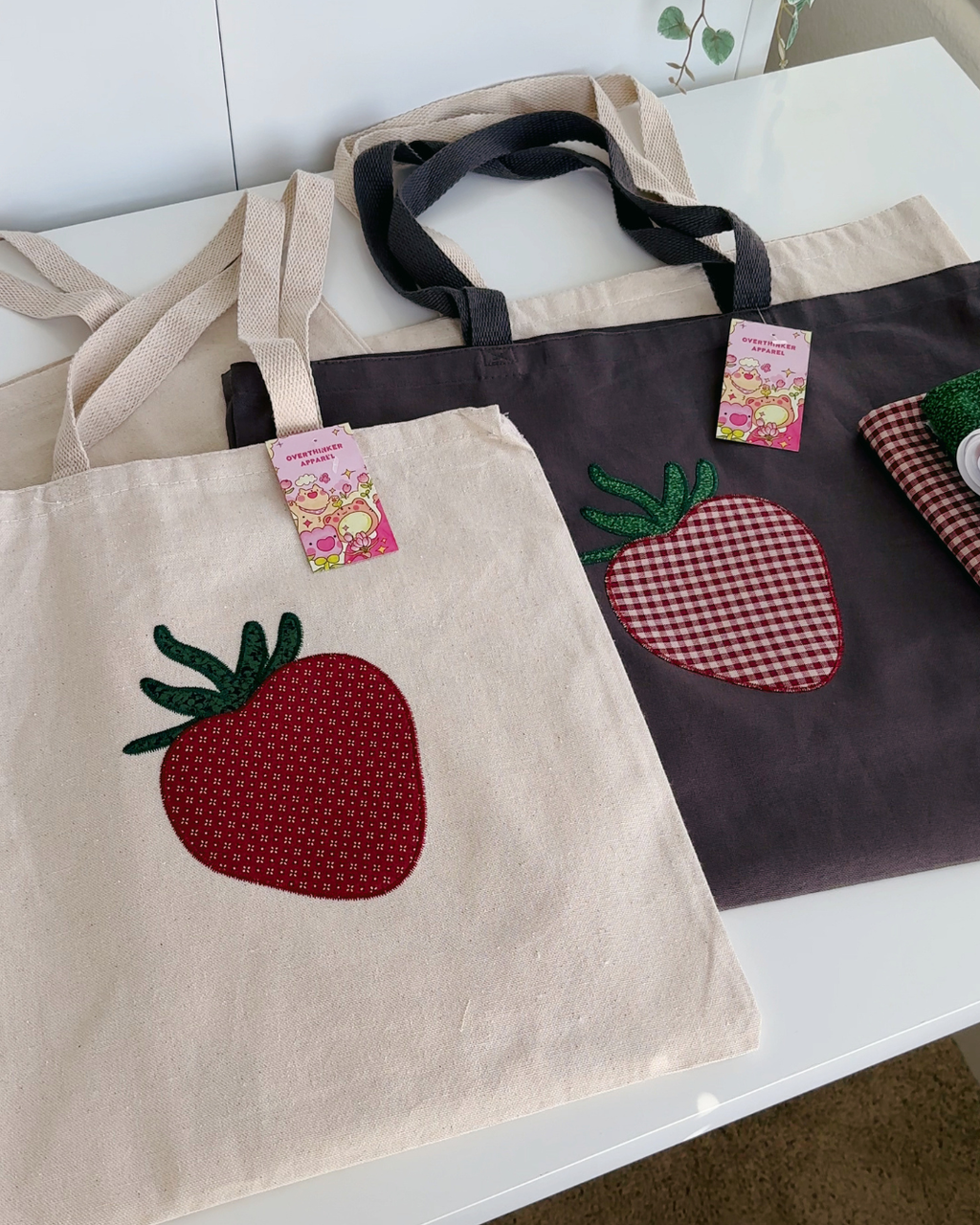 Natural and charcoal canvas tote bags with strawberry embroidery shown together, reusable cotton totes in two color options