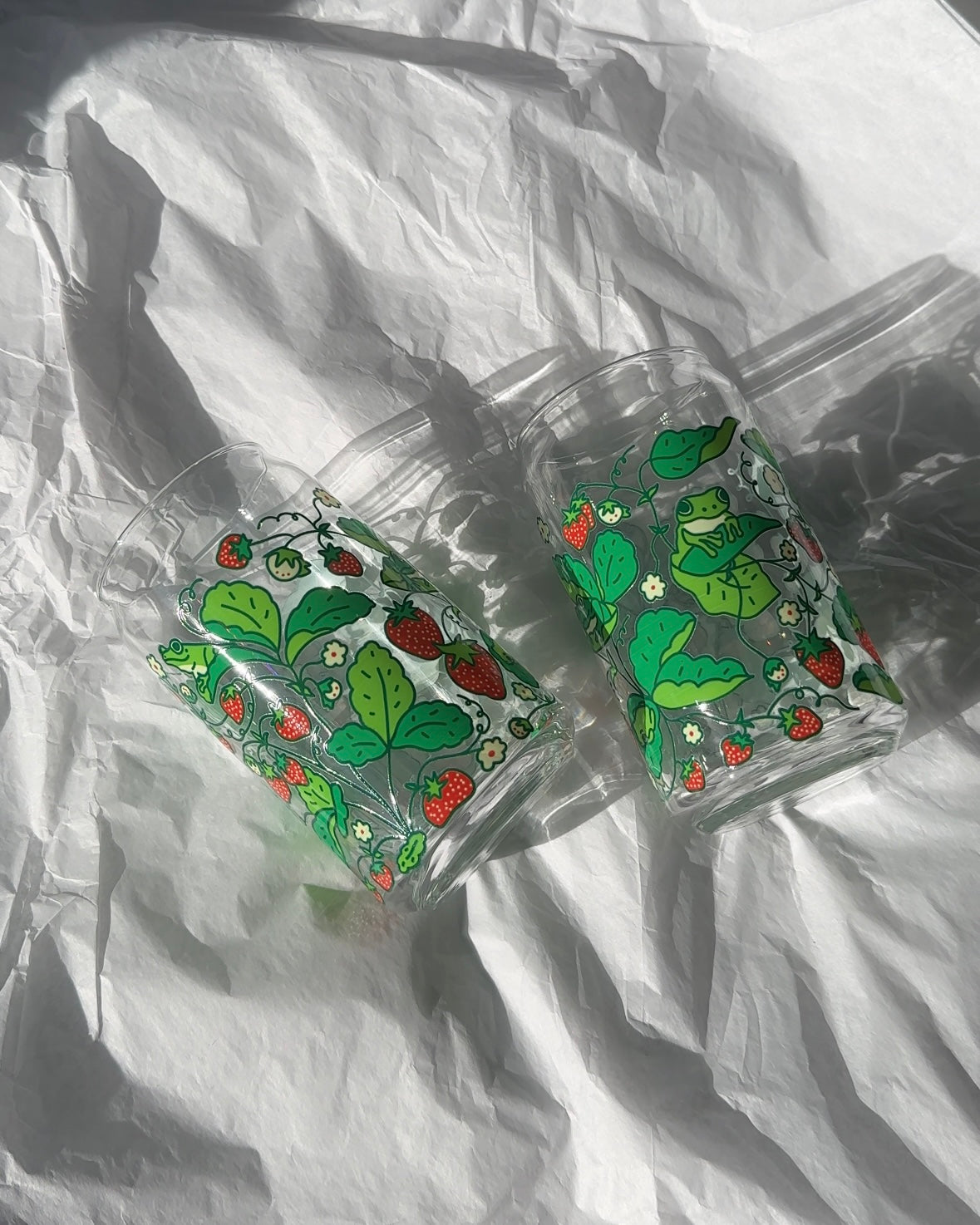 Two clear 16 oz glass cans featuring a cute strawberry frog illustration with green vines and berries, styled together in bright natural light.