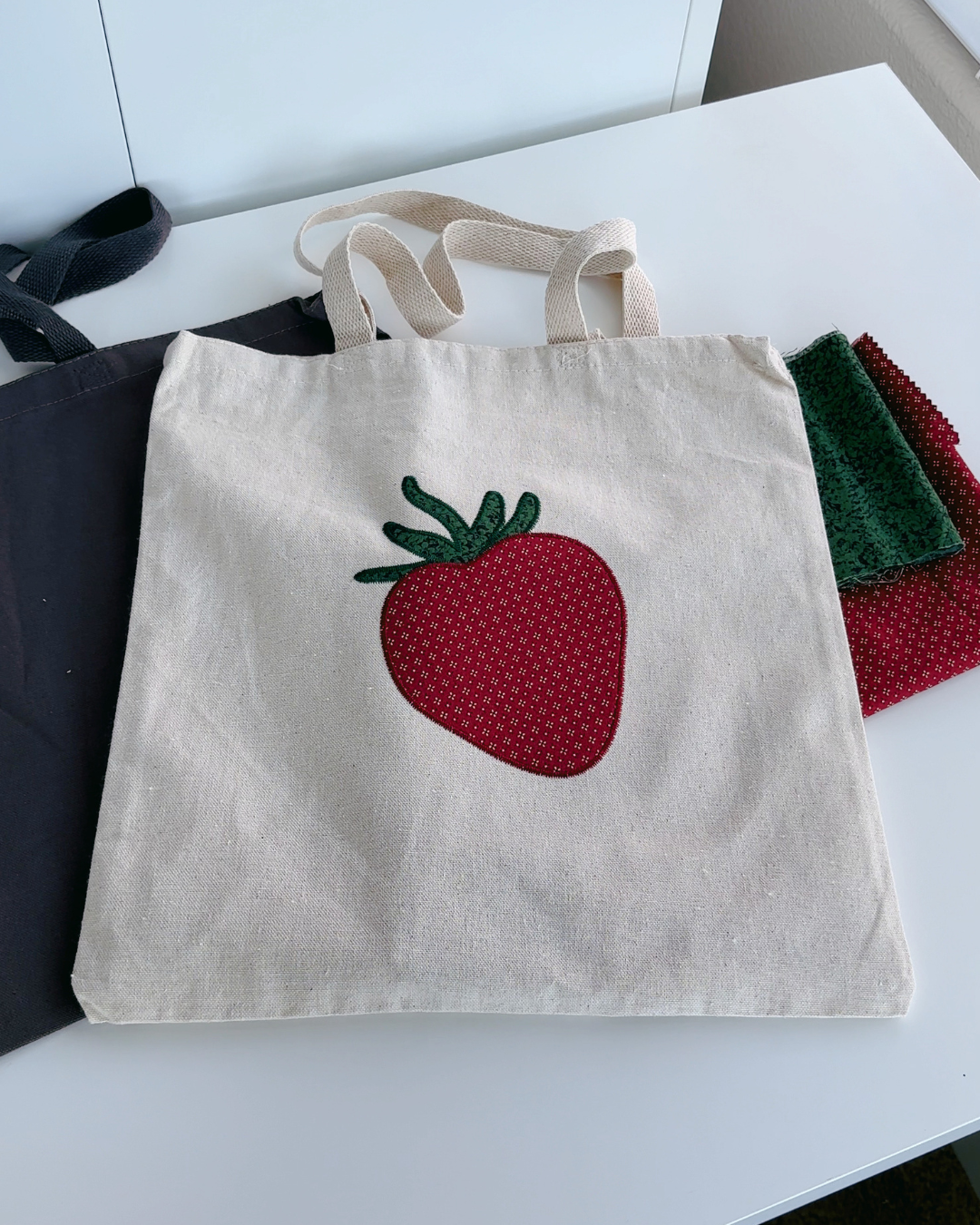 Natural canvas tote with strawberry embroidery next to fabric pieces showing patchwork gingham material and handmade design process