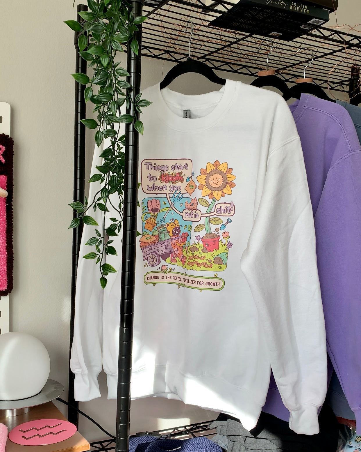 White unisex crewneck sweatshirt hanging on a rack, featuring a detailed garden illustration and an encouraging growth message about progress, change, and putting in the work.