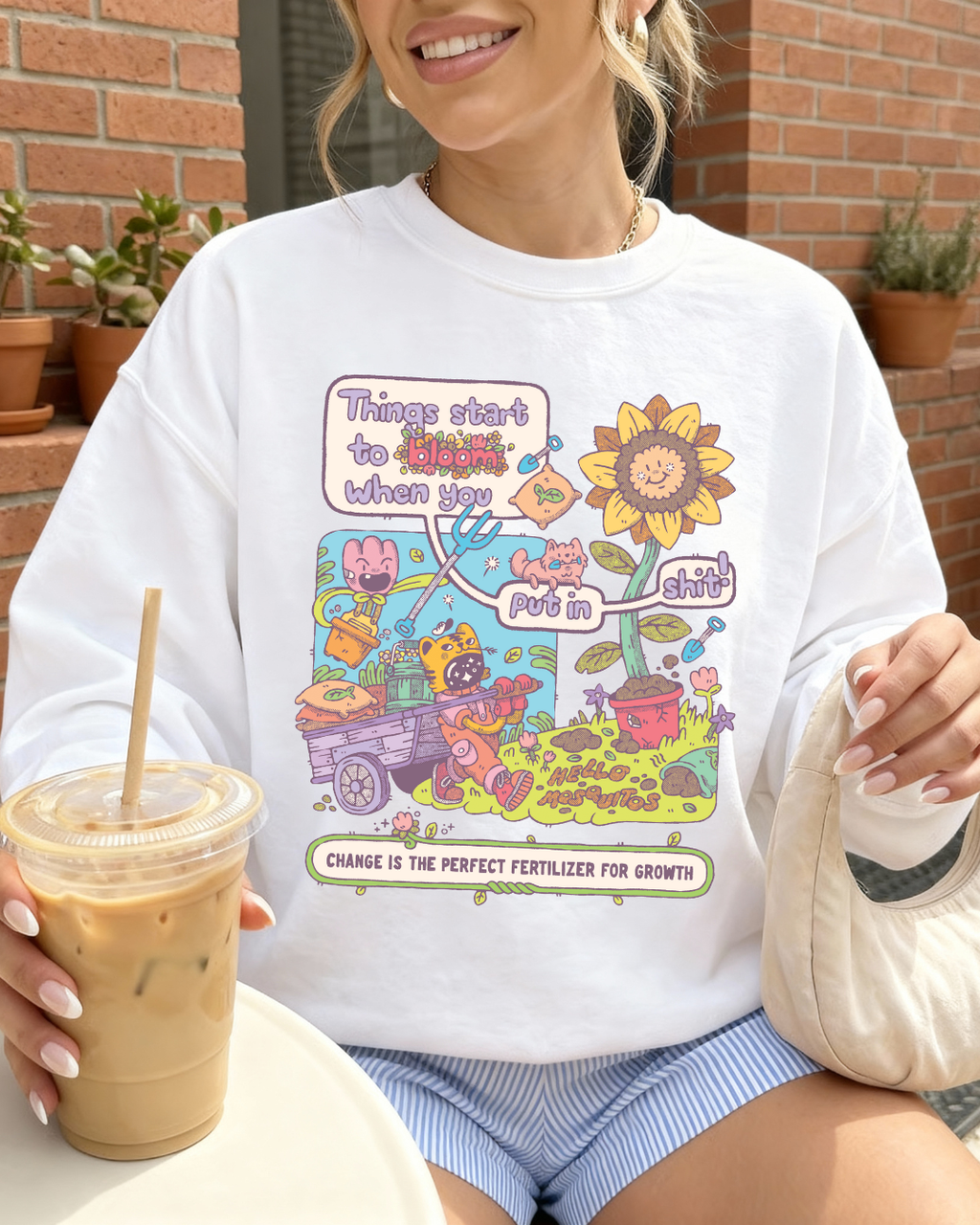 White crewneck sweatshirt with a colorful garden illustration and the phrase "Things start to bloom when you put in the work," styled casually with iced coffee for an encouraging growth mindset look
