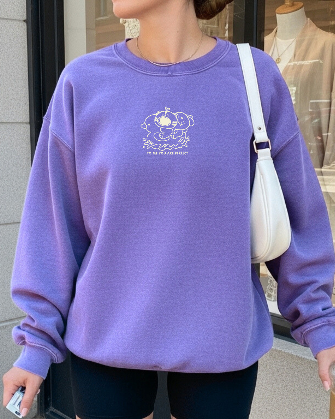 Model wearing a violet embroidered affirmation crewneck sweatshirt with a small white design reading “To me, you are perfect,” featuring two cute characters and a calm, comforting aesthetic.