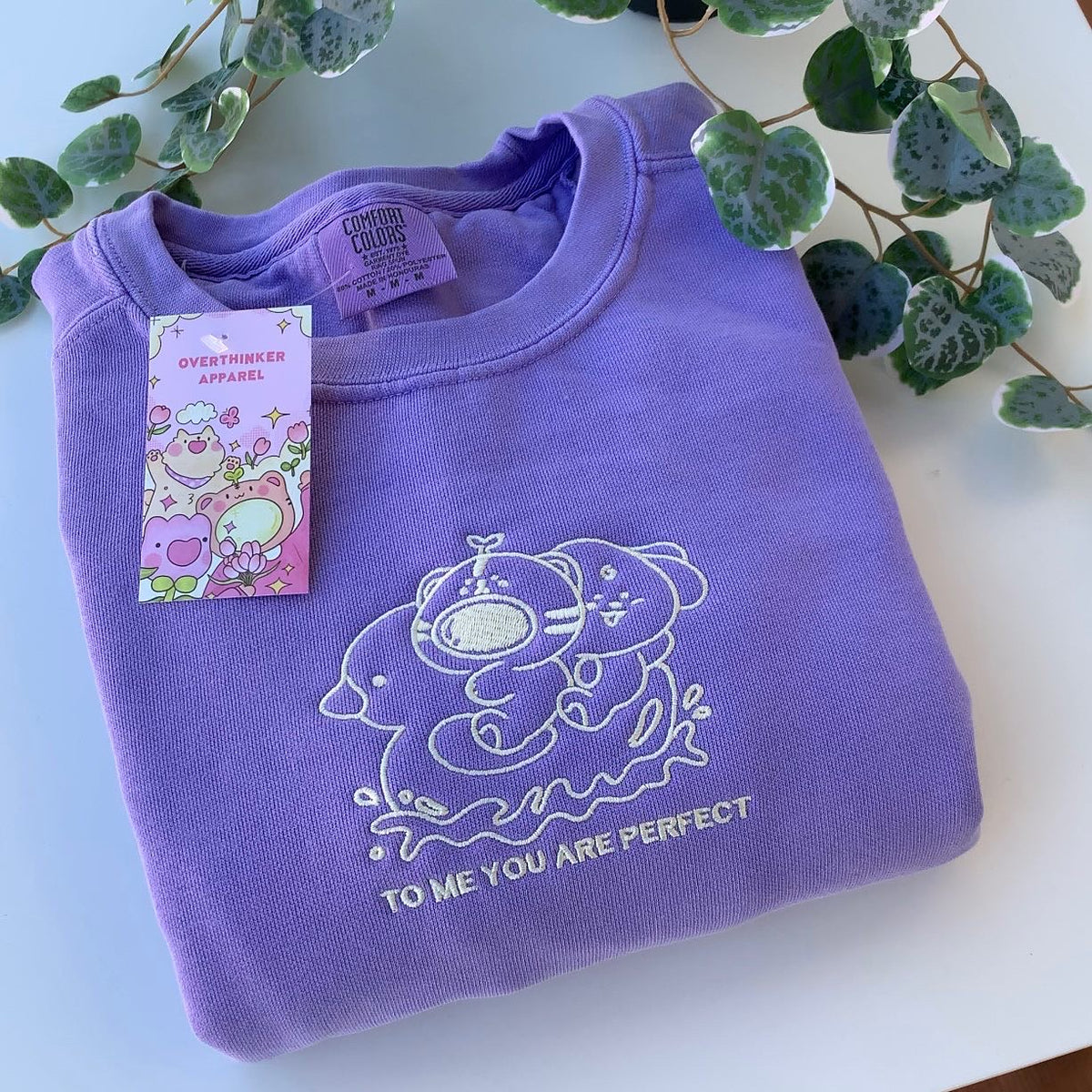 Folded violet embroidered sweatshirt with a soft white character design and the phrase ‘To me, you are perfect,’ shown in a calm flat lay with greenery.