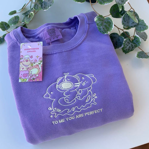 Folded violet embroidered sweatshirt with a soft white character design and the phrase ‘To me, you are perfect,’ shown in a calm flat lay with greenery.