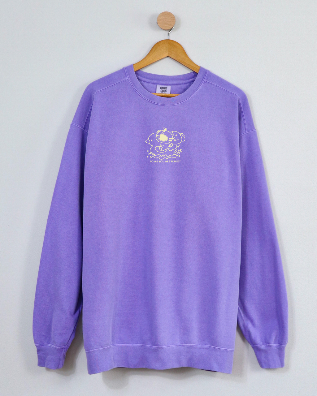 Violet embroidered crewneck sweatshirt hanging on a wooden hanger, featuring two characters and the phrase “To Me, You Are Perfect.”