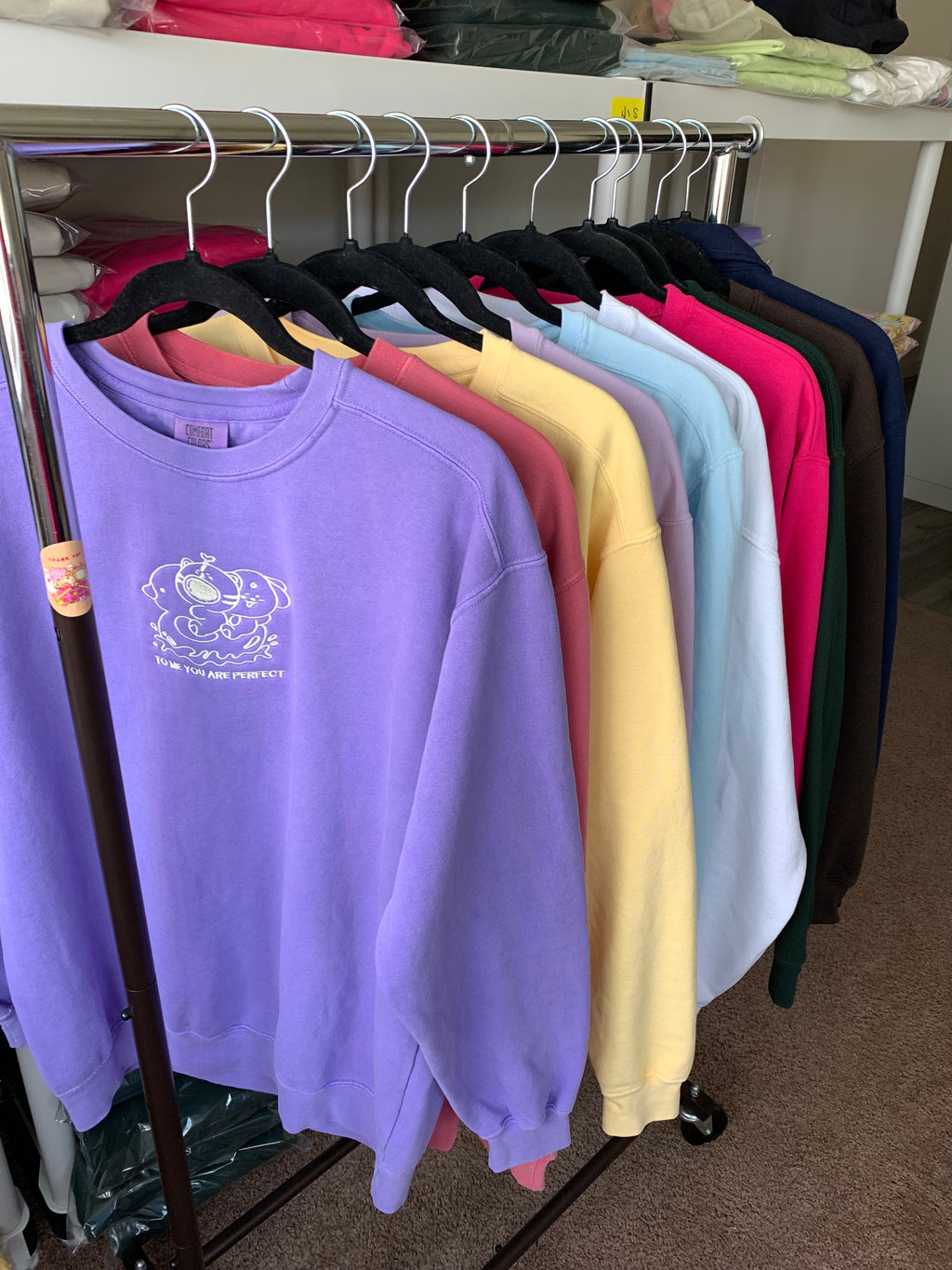 Garment-dyed embroidered affirmation sweatshirts hanging on a rack, showing multiple soft color options including violet, yellow, blue, and pink.