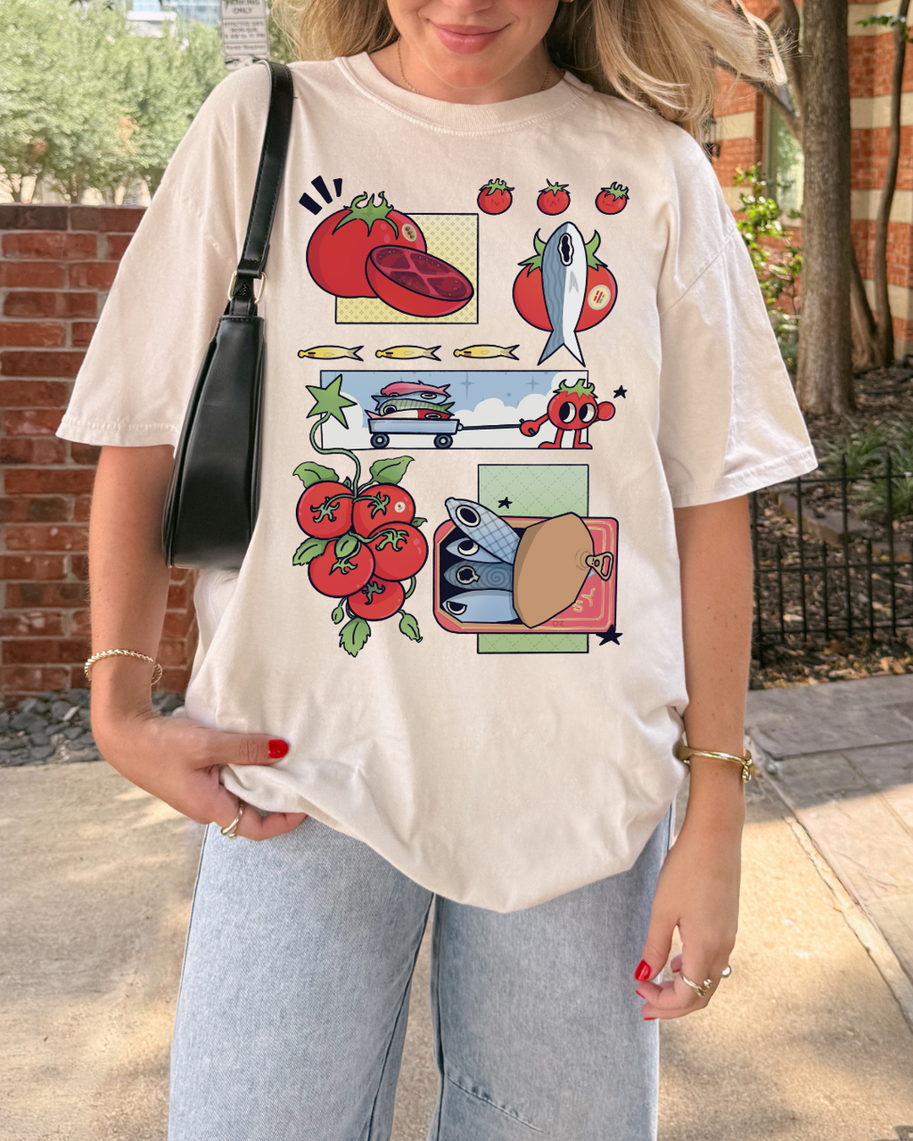 Person wearing an ivory graphic t-shirt with comic-style illustrations of tomatoes and sardines outdoors