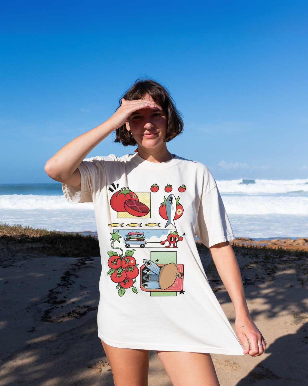 Person wearing an ivory graphic tee with illustrated tomatoes and sardines standing near the ocean