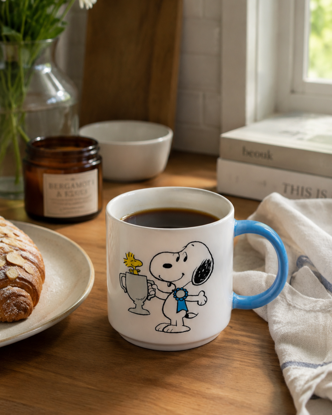 A blue-handled Snoopy mug featuring Snoopy holding a trophy with Woodstock on top sits on a kitchen counter with a croissant, candle, and stacked books.