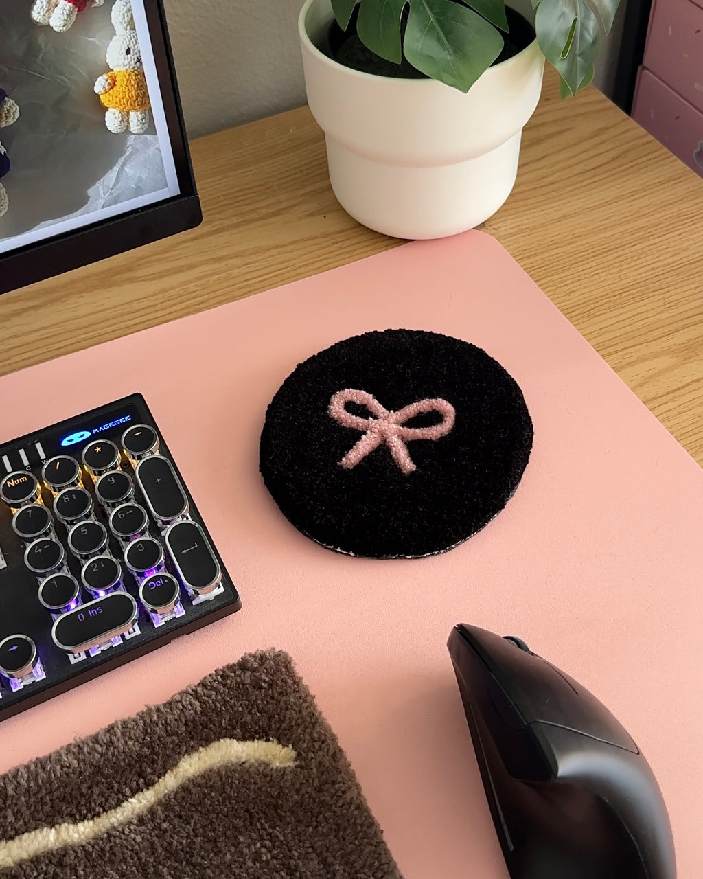 Black tufted bow coaster on pink desk mat beside computer and plant