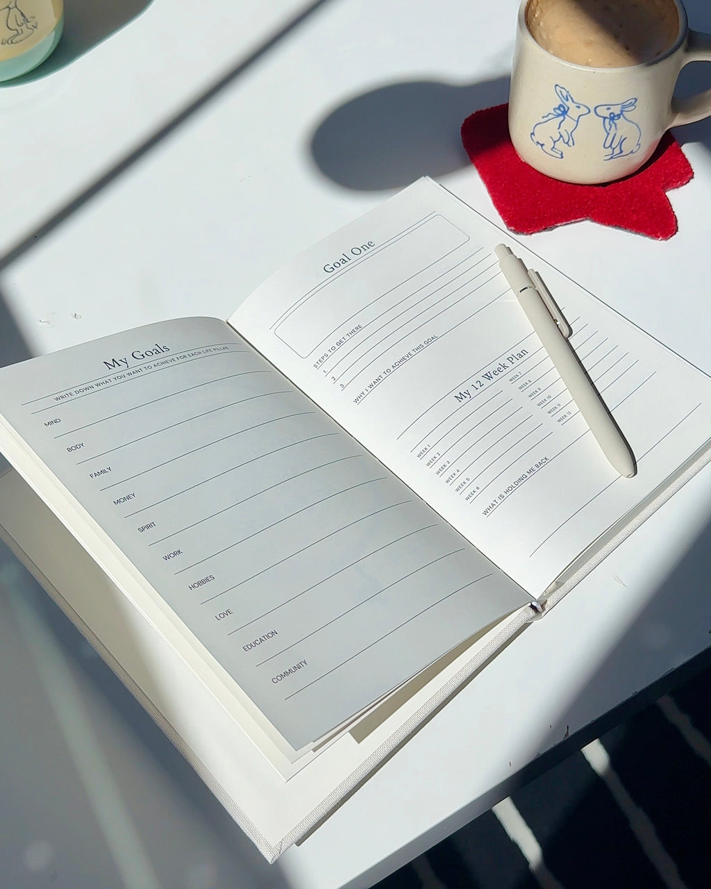 Undated daily planner open to goal planning pages on a white desk with pen and coffee, minimal linen planner for mindful productivity and gentle planning.