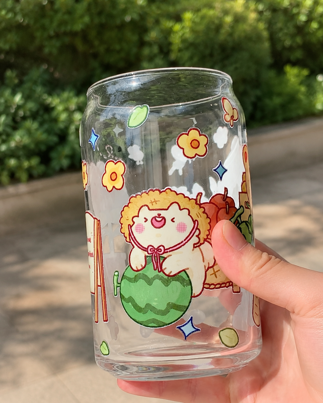 A clear glass can-shaped tumbler with a cute cartoon hedgehog illustration holding a watermelon, held by a hand outdoors with a blurred green garden background.