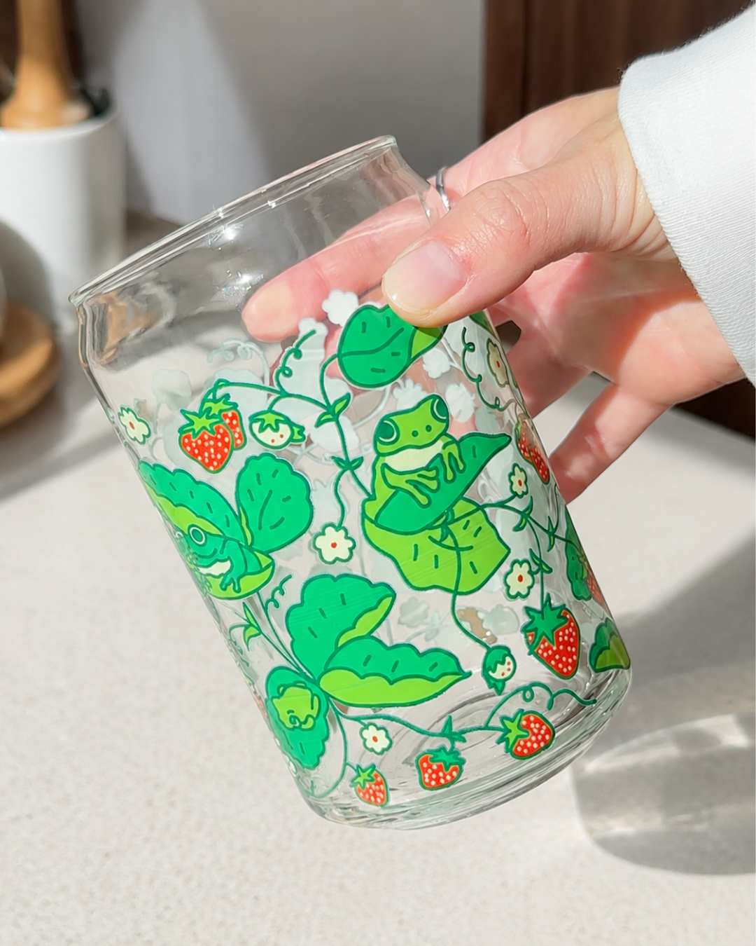 A hand holding a clear glass cup decorated with a green frog, strawberry, and leaf pattern, set against a bright kitchen background.