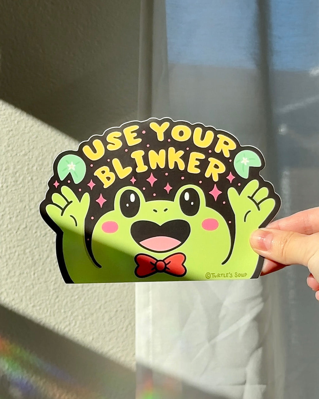 Hand holding a funny vinyl car sticker with a green frog and the words “Use Your Blinker,” bold high-contrast design for car windows or accessories.