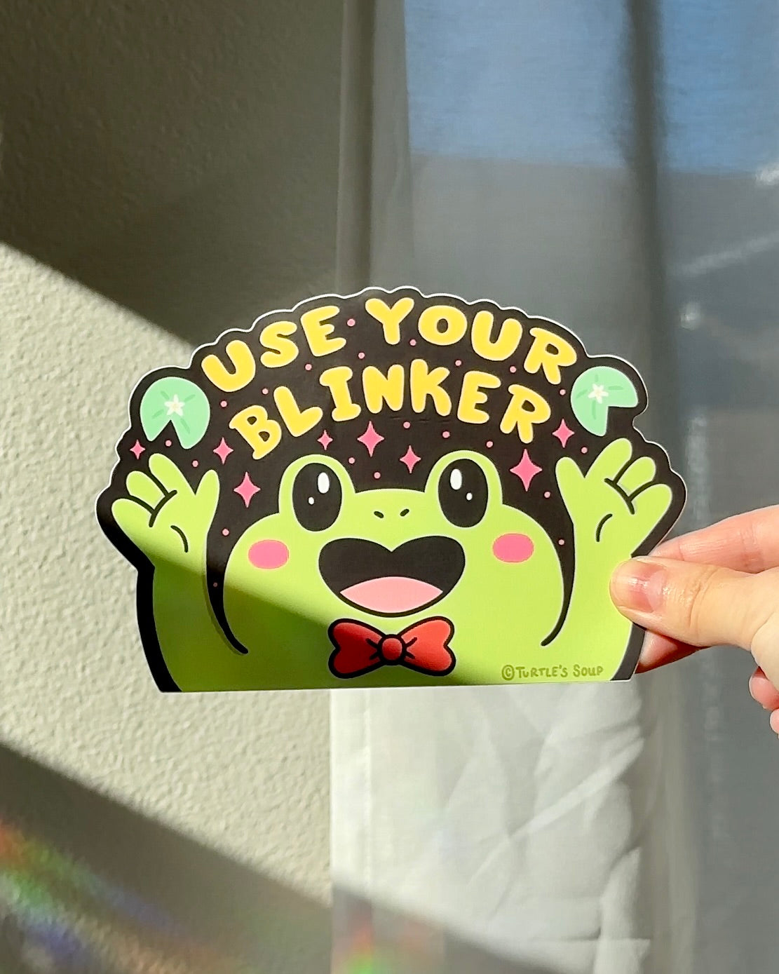 Hand holding a funny vinyl car sticker with a green frog and the words “Use Your Blinker,” bold high-contrast design for car windows or accessories.