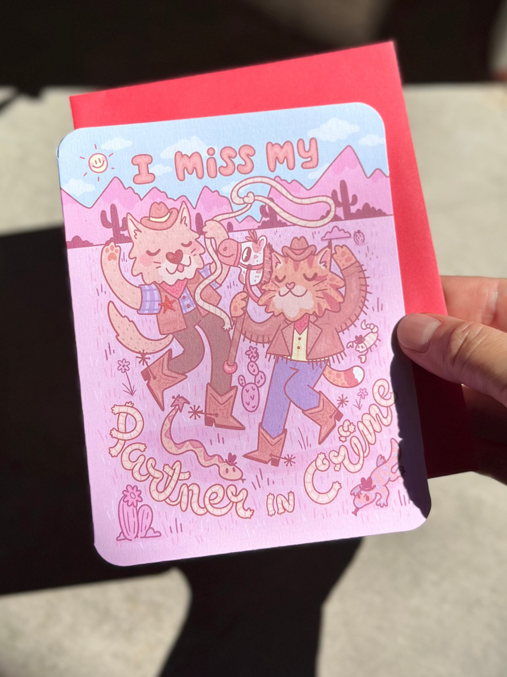 Pink western-themed greeting card with two cartoon cowboy cats dancing in the desert, wearing boots and hats, surrounded by cacti and snakes, with text ‘I miss my partner in crime,’ paired with red envelope.